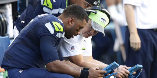 Russell Wilson and a coach sit on the sidelines of a preseason game looking at a Surface tablet.