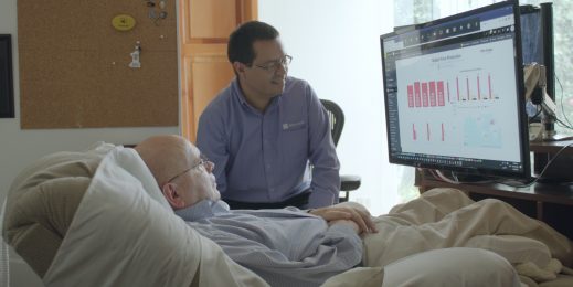 One man lies in a bed and another sits at his side, both looking at a computer screen