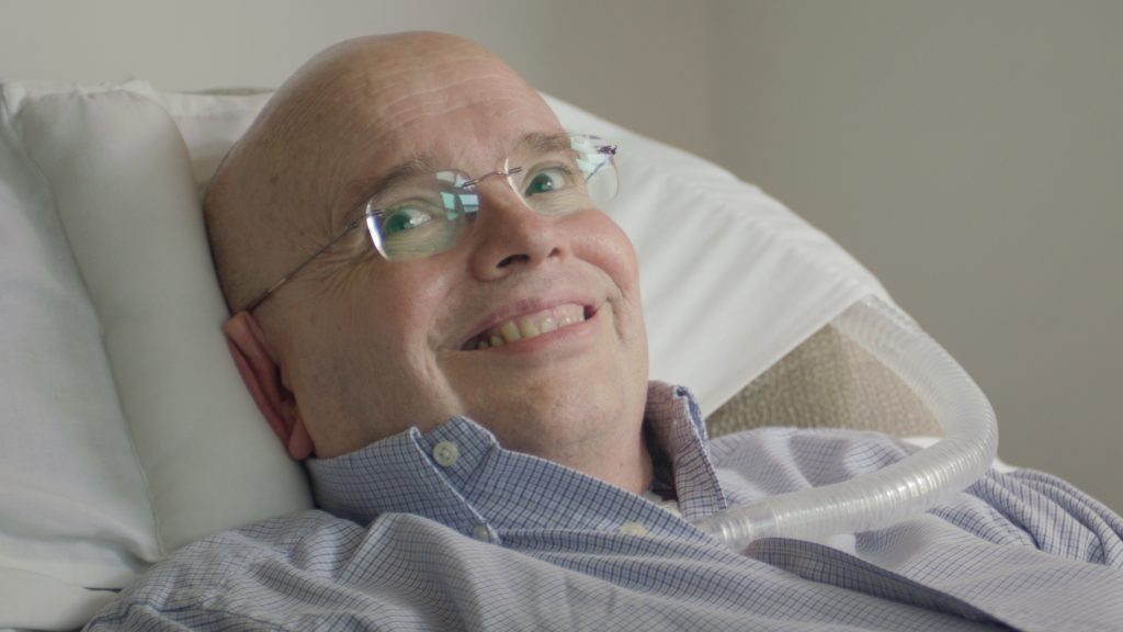 A white man with a bald head, wearing glasses, has a big smile as he leans against a pillow