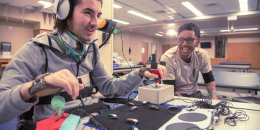 A man using a wheelchair tries out adaptive controllers to play a video game as another man watches and grins