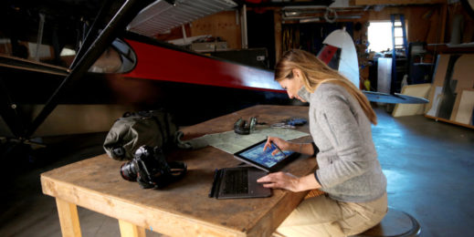 Woman with long hair sitting at a drafting table, using a digital pen, with a small aircraft in the background