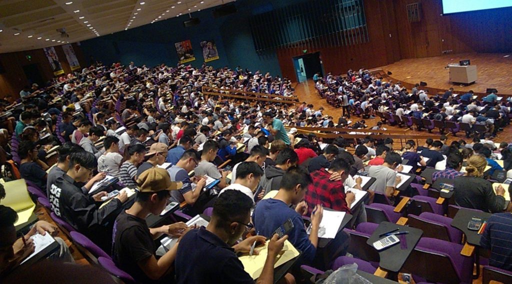 Hundreds of students sit in a large lecture hall, looking down at papers on their desks.
