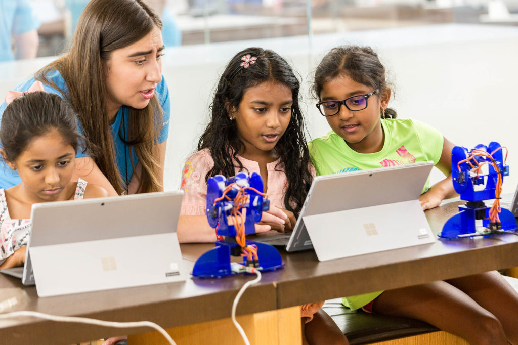 Three girls look intently at laptops near small robots while a female Microsoft Store employee helps them
