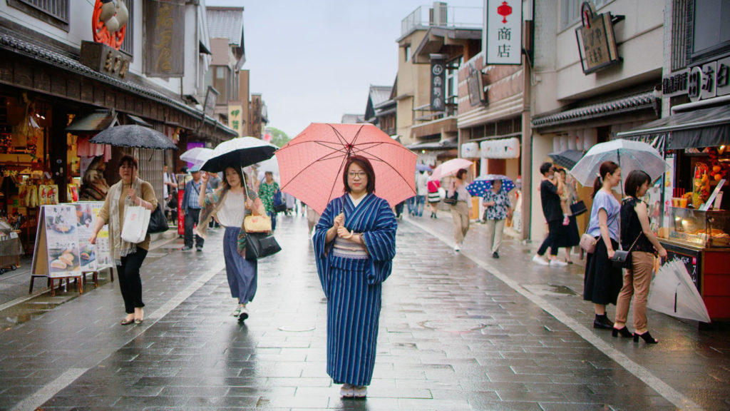 A woman wearing traditional Japanese clothing stands on a city street in Japan holding an umbrella