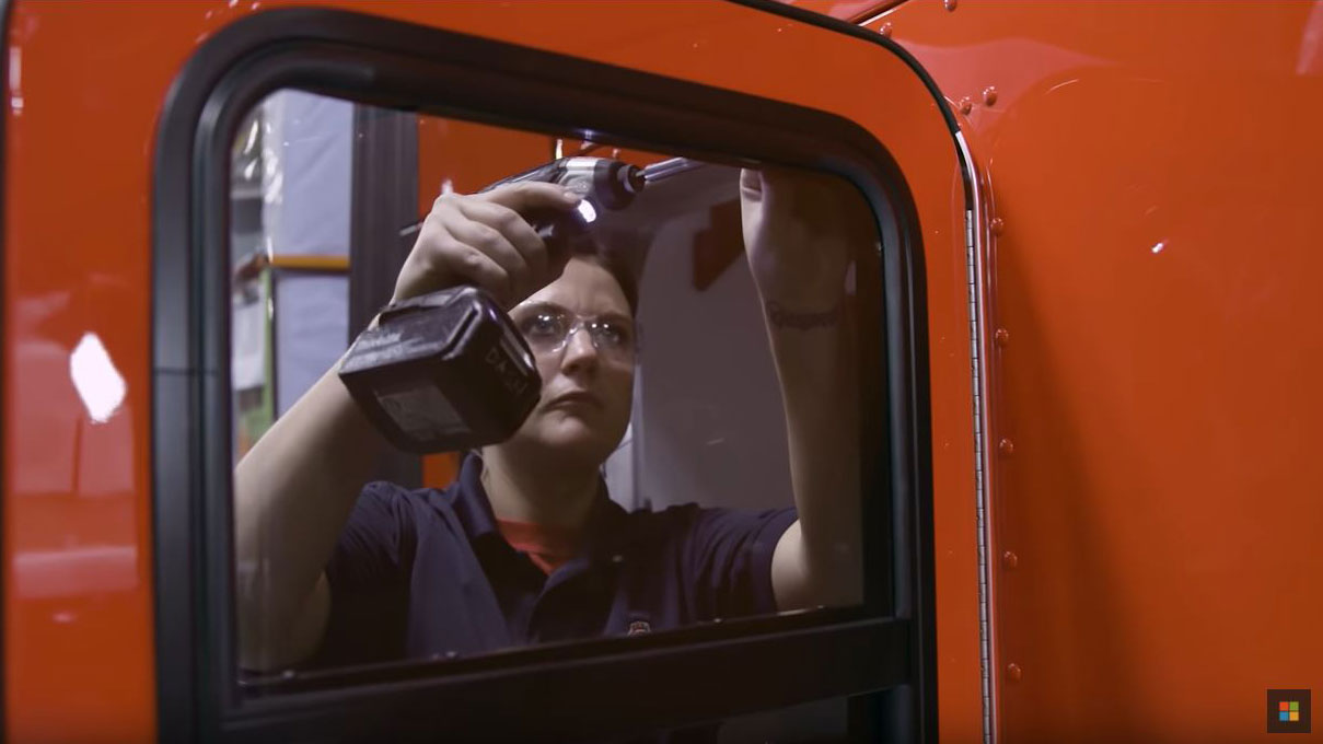 A woman uses a drill to work on the door frame of a large vehicle