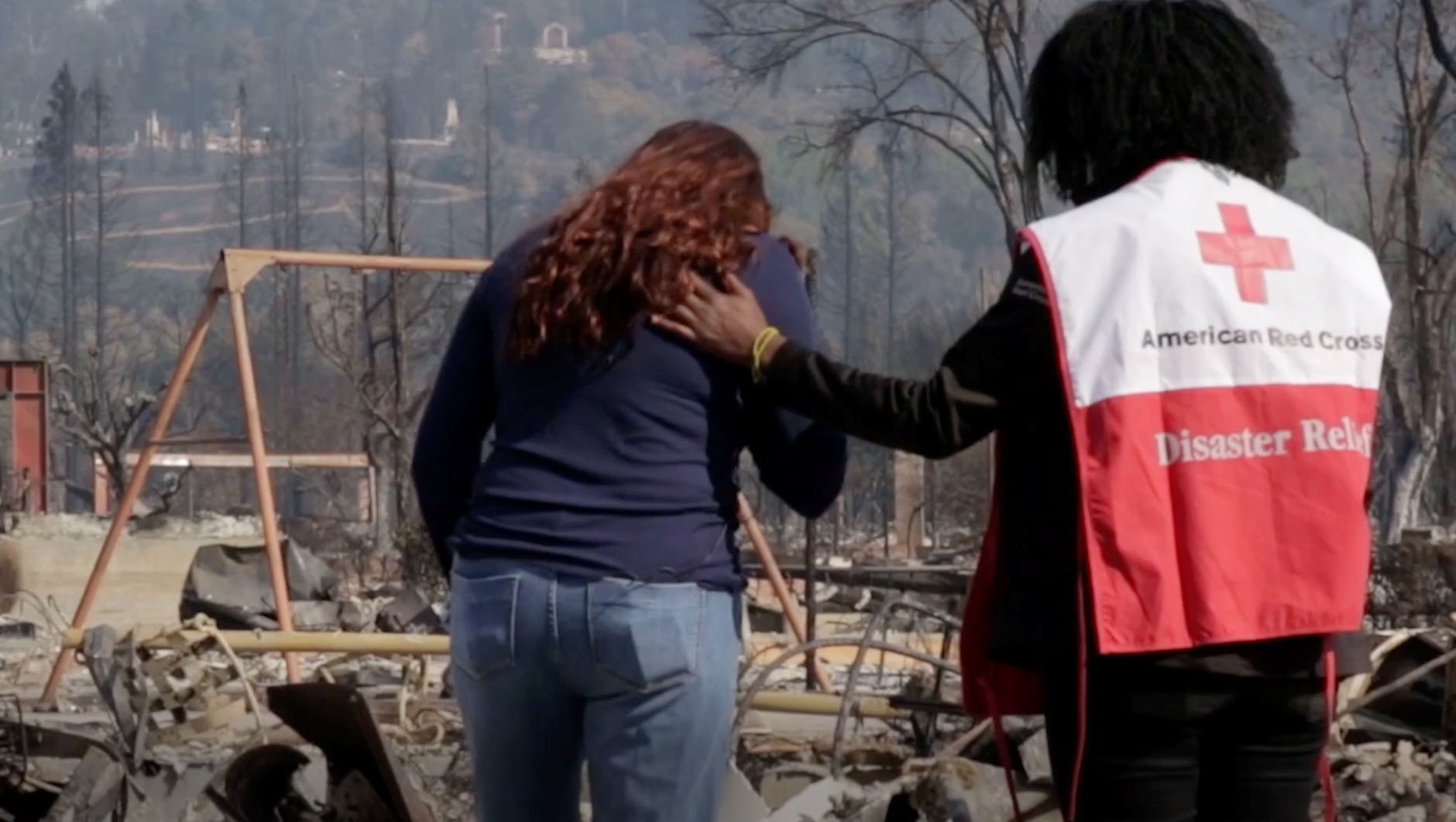 A volunteer wearing a Red Cross vest comforts someone at the scene of a disaster