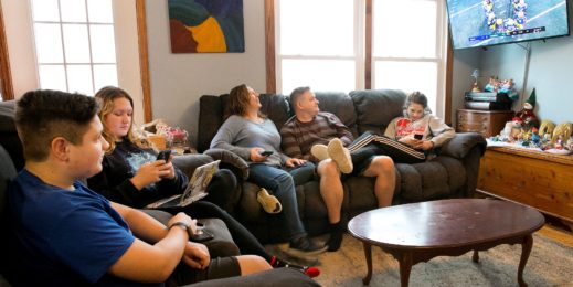 A family sits together on a large sectional couch in their home, some looking at tablets and devices while others watch a football game on TV