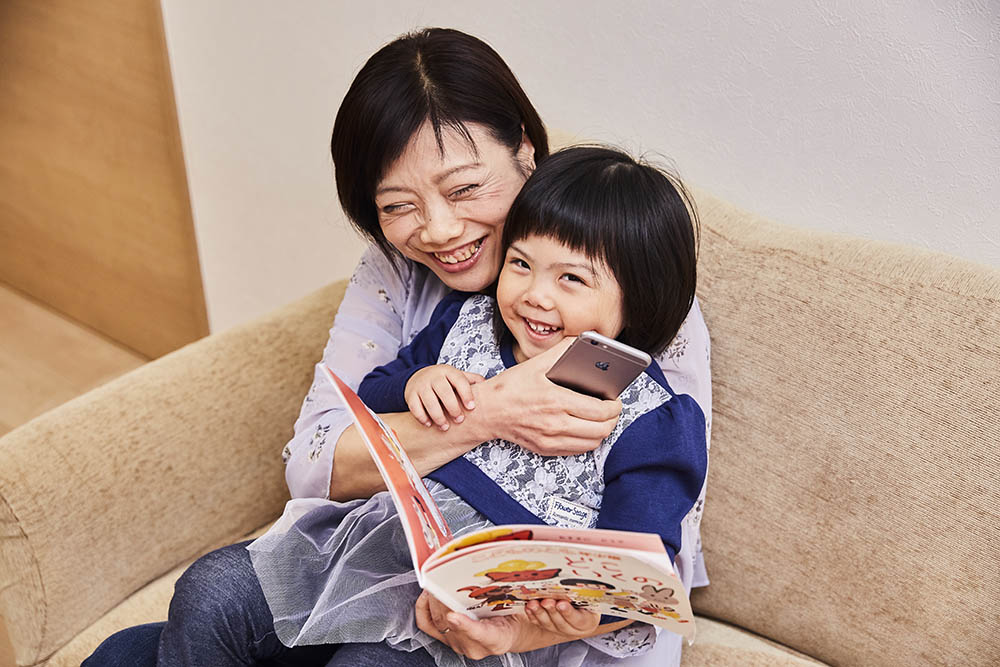 A mother smiles and hugs her young daughter while holding a phone and book 