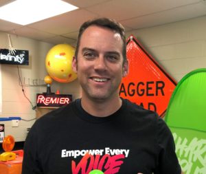 Smiling man stands in a classroom filled with colorful decor and signage