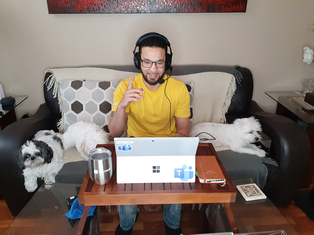 Carl Richardson smiles and wears a headset and yellow Microsoft Store shirt while working on his couch next to his three small dogs