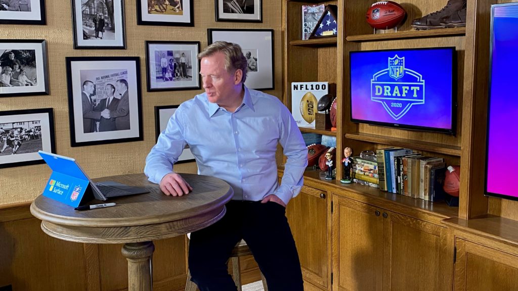 Man sits at a table in front of his Surface device with large screens that say NFL draft and various sports-themed decor in the background