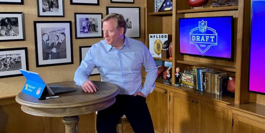 Man sits at a table in front of his Surface device with large screens that say NFL draft and various sports-themed decor in the background