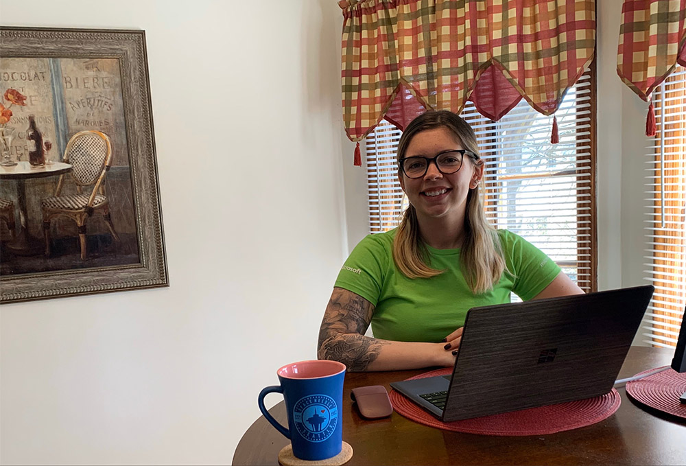 Samantha Janiec wears a green Microsoft Store shirt and smiles in front of her laptop while working from home