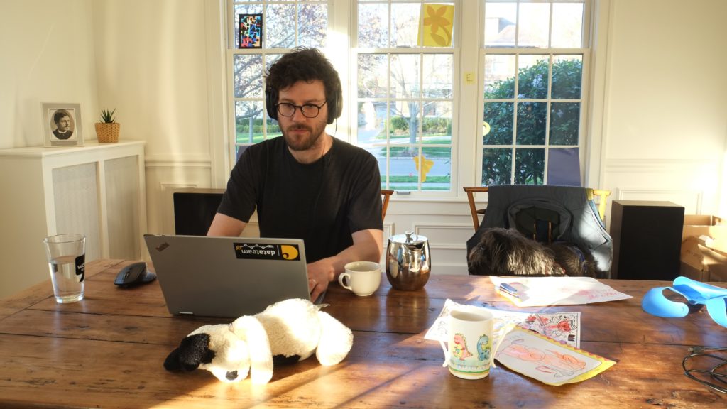 man sits in front of laptop at table with dog, toys and coffee cup around him