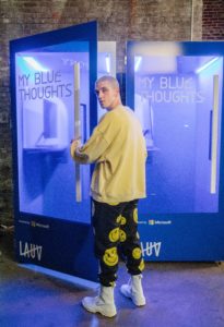 Lauv stands in front of a blue phone booth that reads My Blue Thoughts on the door. Inside a computer is visible. Lauv is looking over his left shoulder, wearing a yellow sweatshirt and black sweats with yellow smiley faces.
