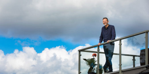 Chuck Edward stands on a deck with blue sky and clouds behind him