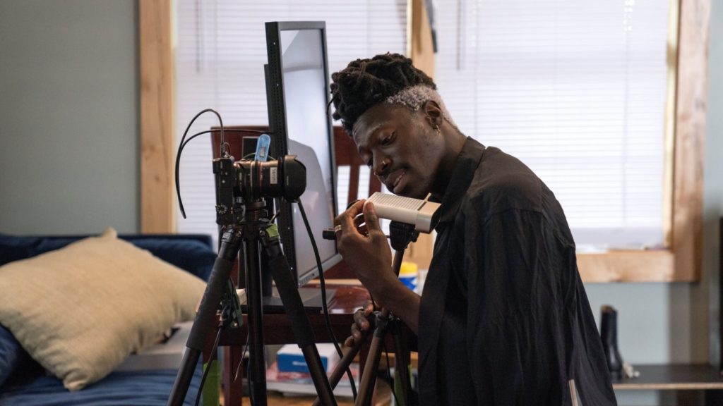 Musician Moses Sumney kneels to examine the Azure Kinect hardware, whi ch is on a tripod next to a mounted camera and a table with a large computer monitor. A blue sofa is in the background; we are in his living room