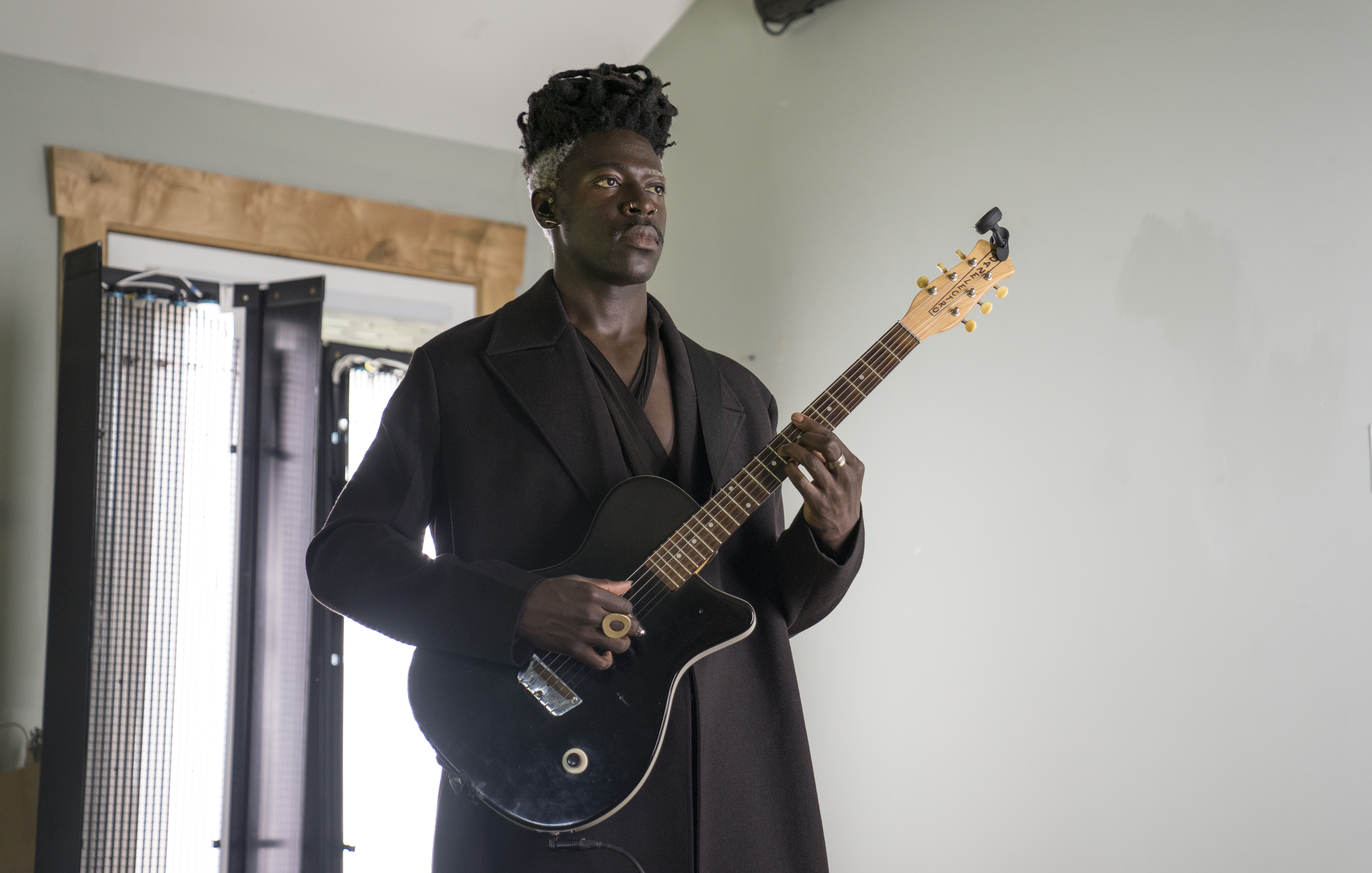 Musician Moses Sumney stands playing a black guitar. He’s wearing a long, dark brown overcoat and has a brass statement ring in the shape of a large O on his right hand.