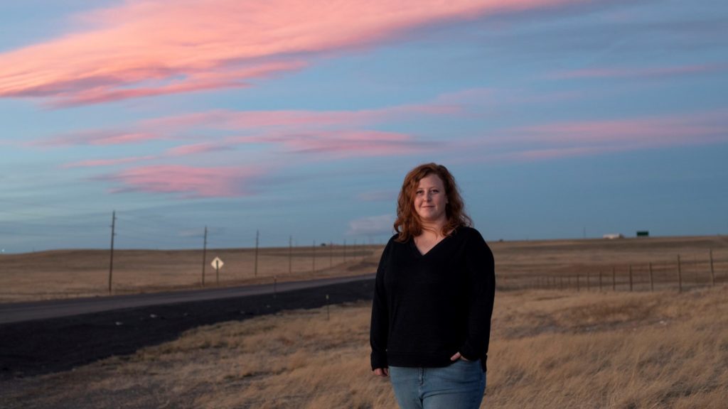 Woman stands near an open field against a blue orange sky during sunset