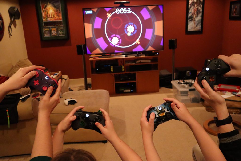 Four pairs of hands holding Xbox controllers in a living room in front of a TV