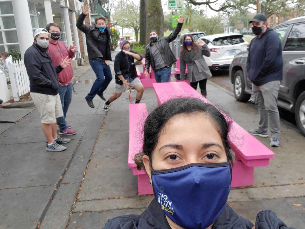 Team shot on a sidewalk, in masks, around a pink picnic table