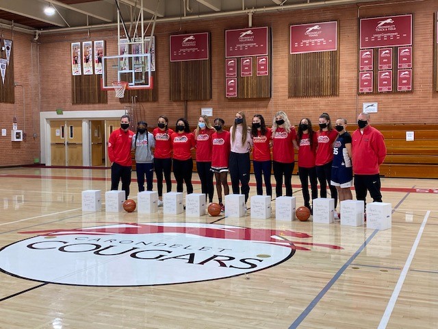 Girls in red shirts and black shorts lined up on a basketball court with the white kits in front of them