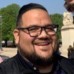 A man with glasses smiles outside on a sunny day