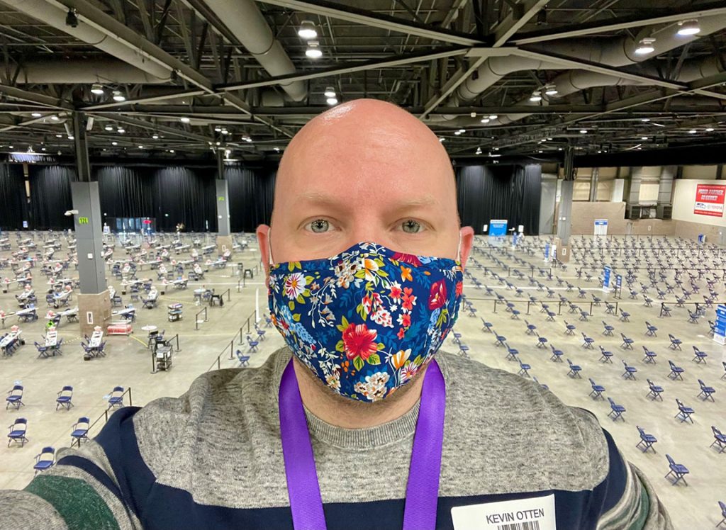Bald man wearing a mask looks into camera, with a large room filled with chairs behind him