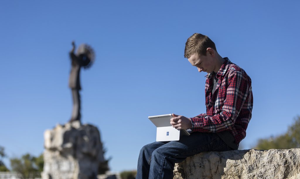 A boy holds a laptop on his lap and sits on a rock near the Keeper of the Plains sculpture
