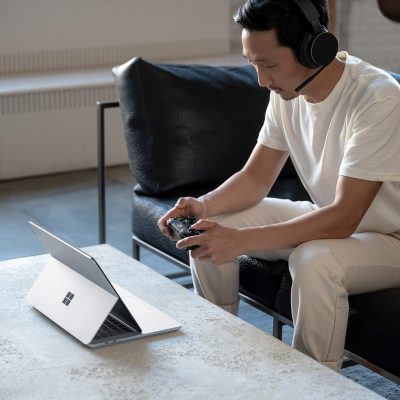 A man sitting down using an Xbox controller with a headset on while on a Surface laptop