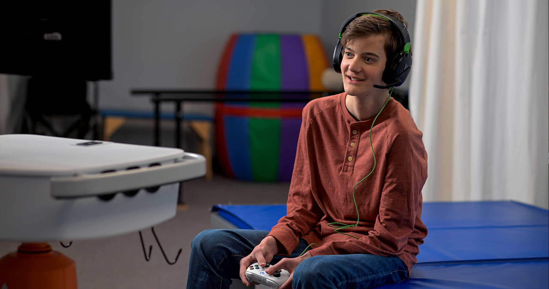 A young man sitting on a bench with a red shirt and headphones, holding a controller in his hands.