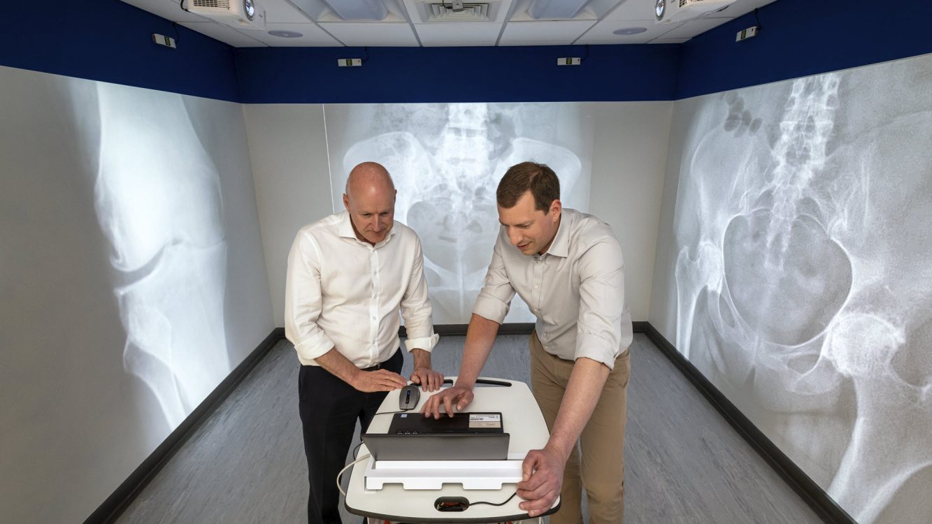 Two men dressed in smart trousers and shirts stand in a small room. They are standing in front of a laptop and are looking at the screen. Large images of knee and hip joint X-rays have been projected onto the walls behind them and either side of them. 