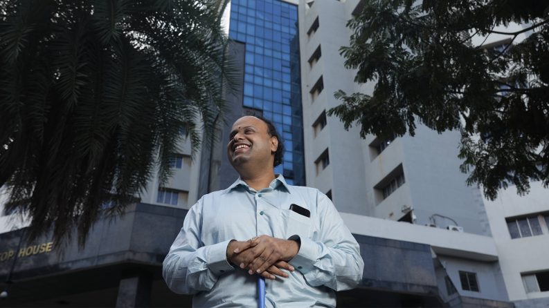 A man who is blind holds his guide cane as he stands in front of an office building