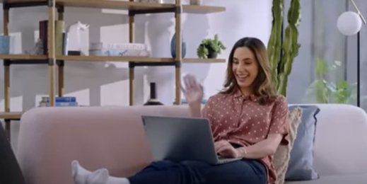Woman sitting on a couch with a laptop computer, waving and smiling at other attendants on a Microsoft Teams meeting