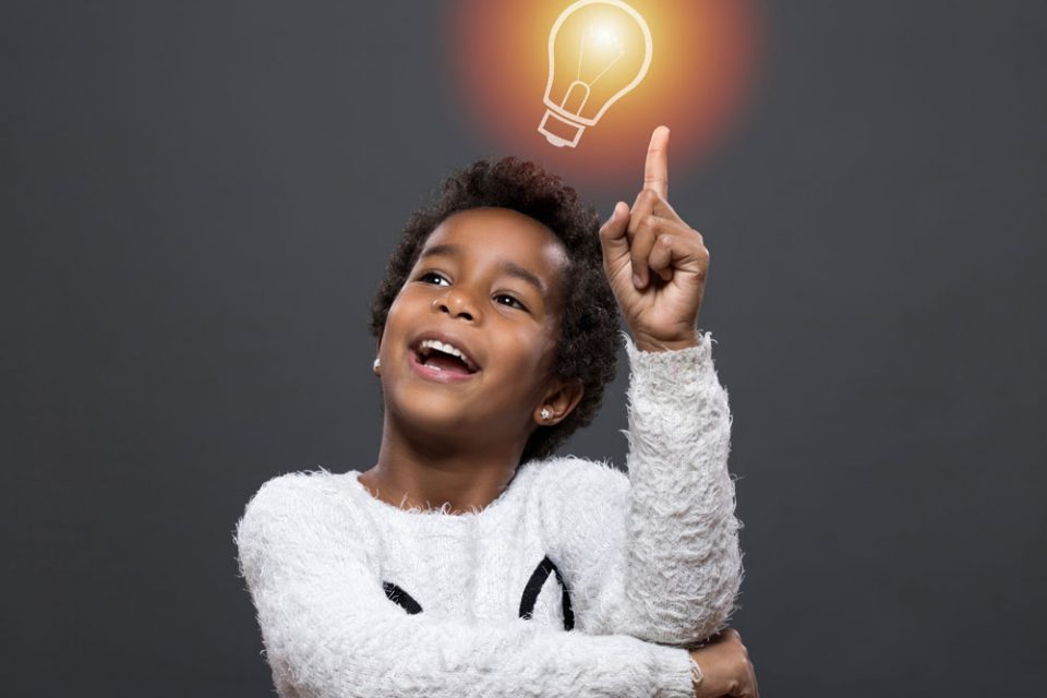 Young girl standing in front of a grey background pointing at a light bulb and smiling