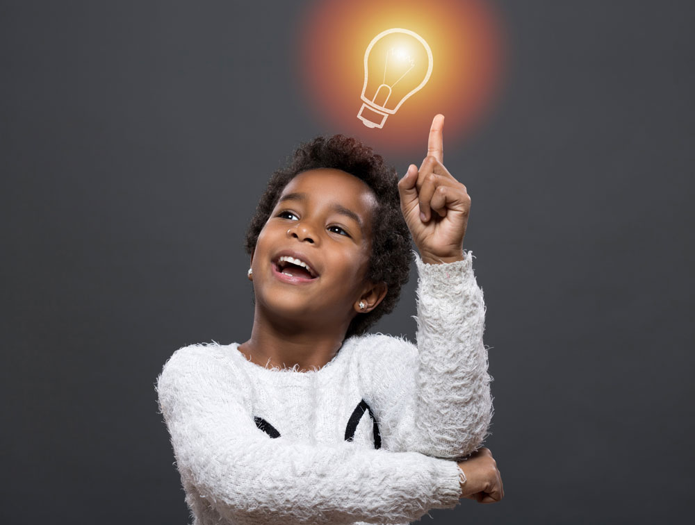 Young girl standing in front of a grey background pointing at a light bulb and smiling