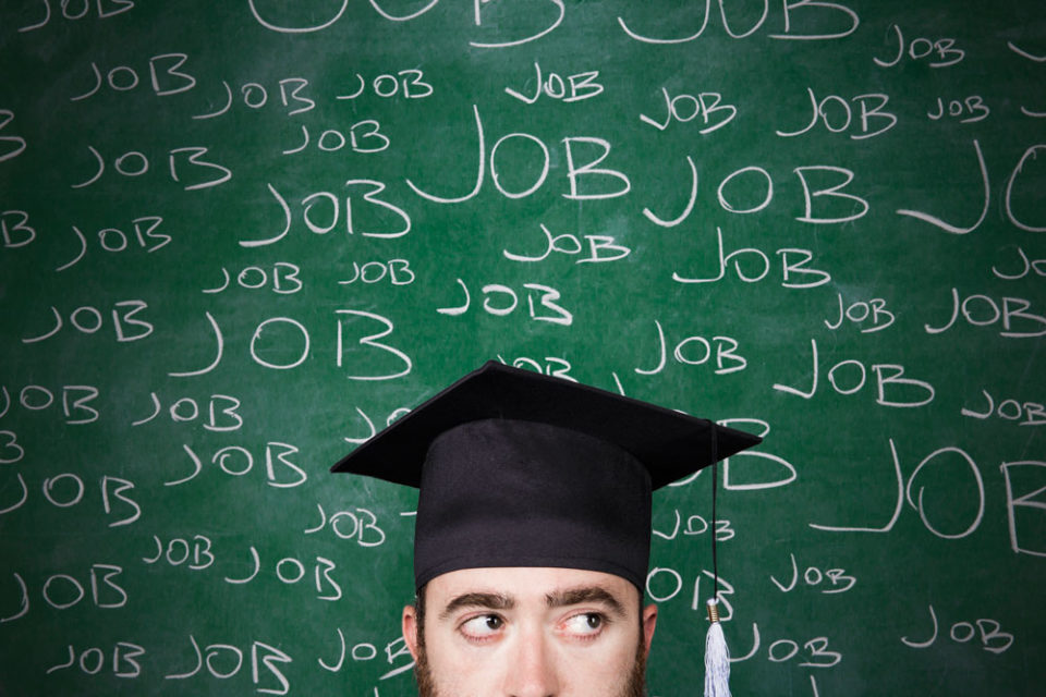 A male wearing a graduation hat in front of a green school board with the word ‘Job’ written on it.