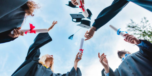 bottom view of happy multicultural graduates with diplomas throwing caps up with blue sky on background