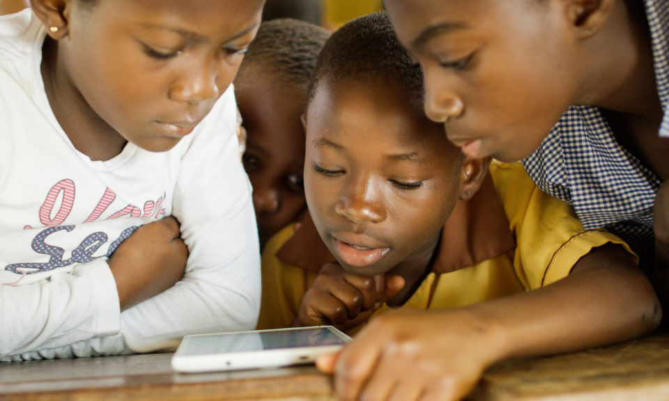 African school children gather around and read from a tablet.