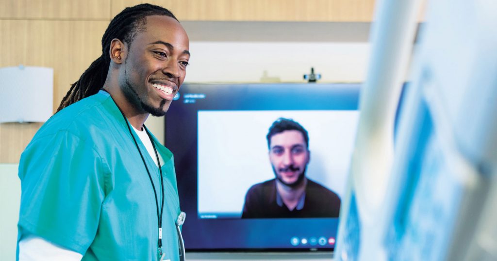 A male doctor is talking to a patient in a hospital bed while a large TV screen behind him displays a video call with another person.