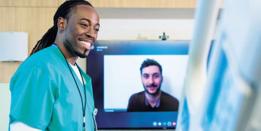 A male doctor is talking to a patient in a hospital bed while a large TV screen behind him displays a video call with another person.