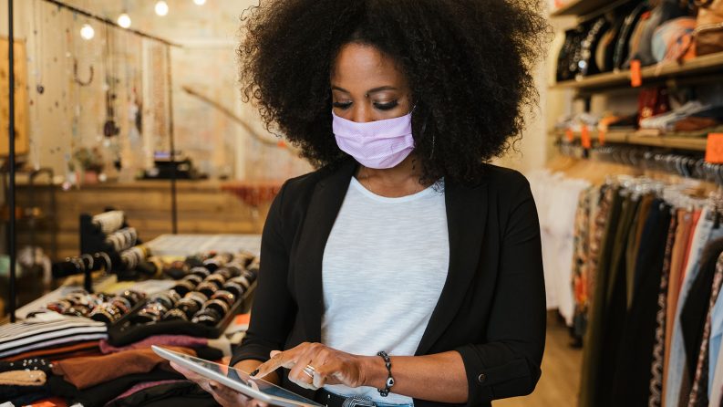 A female shop assistant wearing a face mask checks her tablet in a clothing store