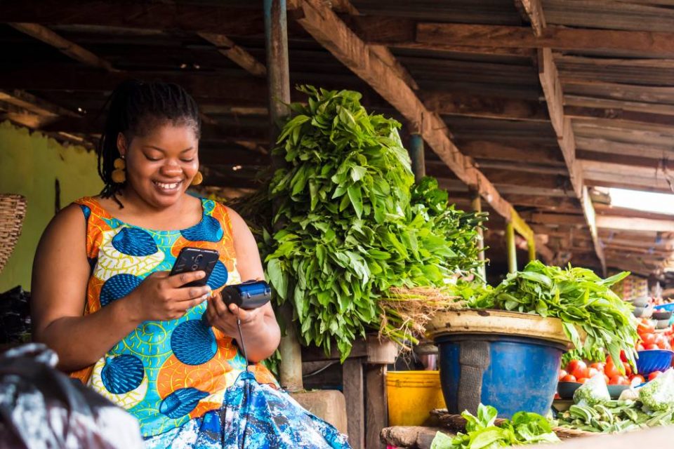 A female business owner smiling at her smartphone and card machine while standing outside at her farm stall of vegetables.