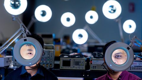 Two men sit behind magnifying classes in a lab setting