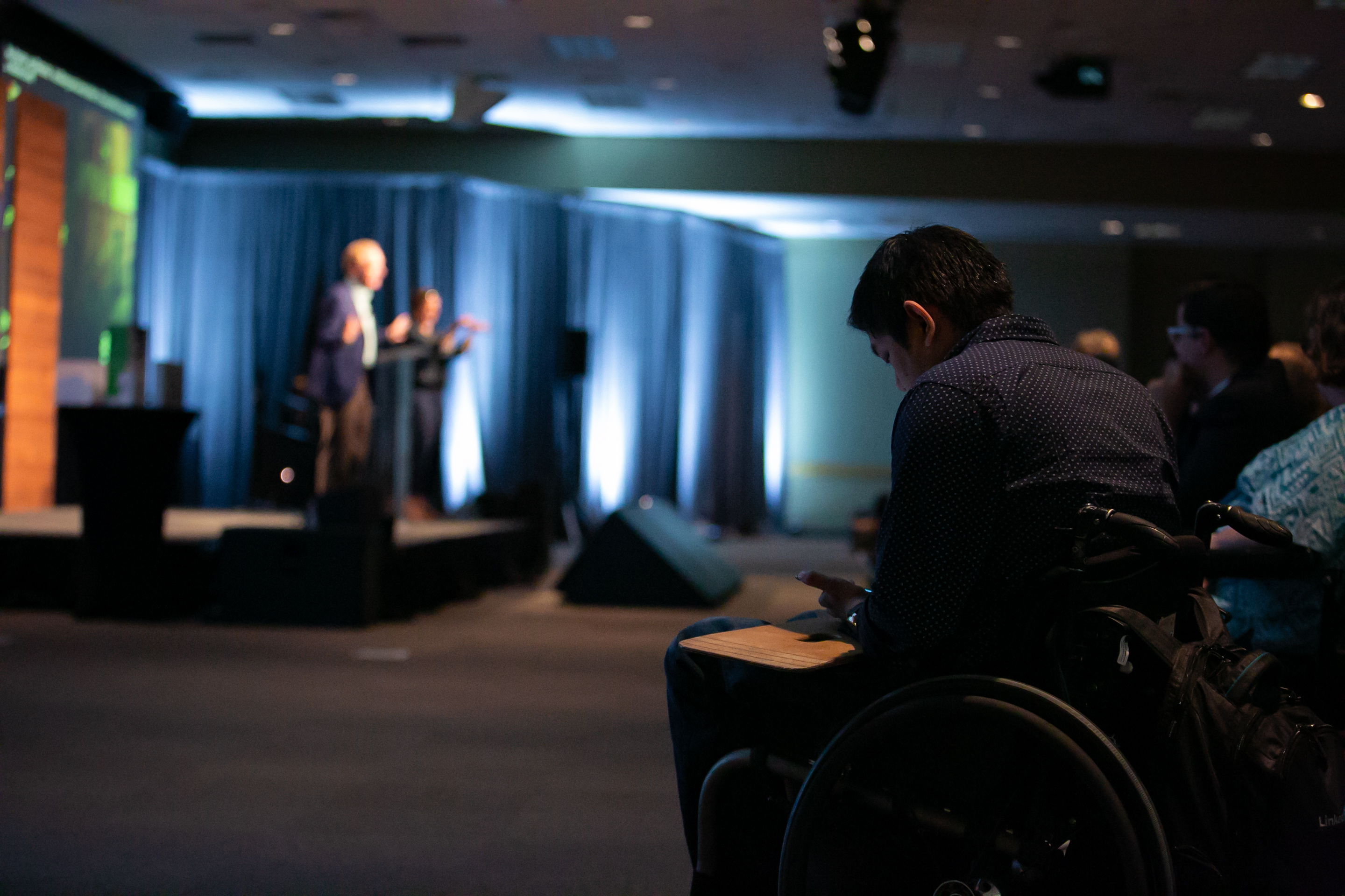Photo of man in the background speaking on stage while a man in a wheelchair takes notes in the audience.