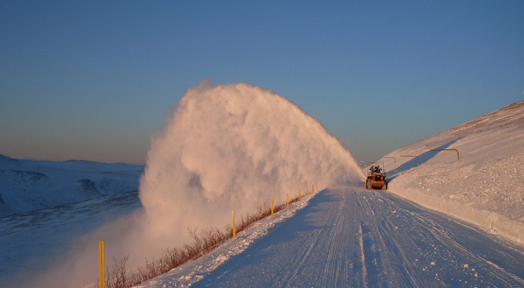 Machine blows arc of snow off a snowy road against blue sky
