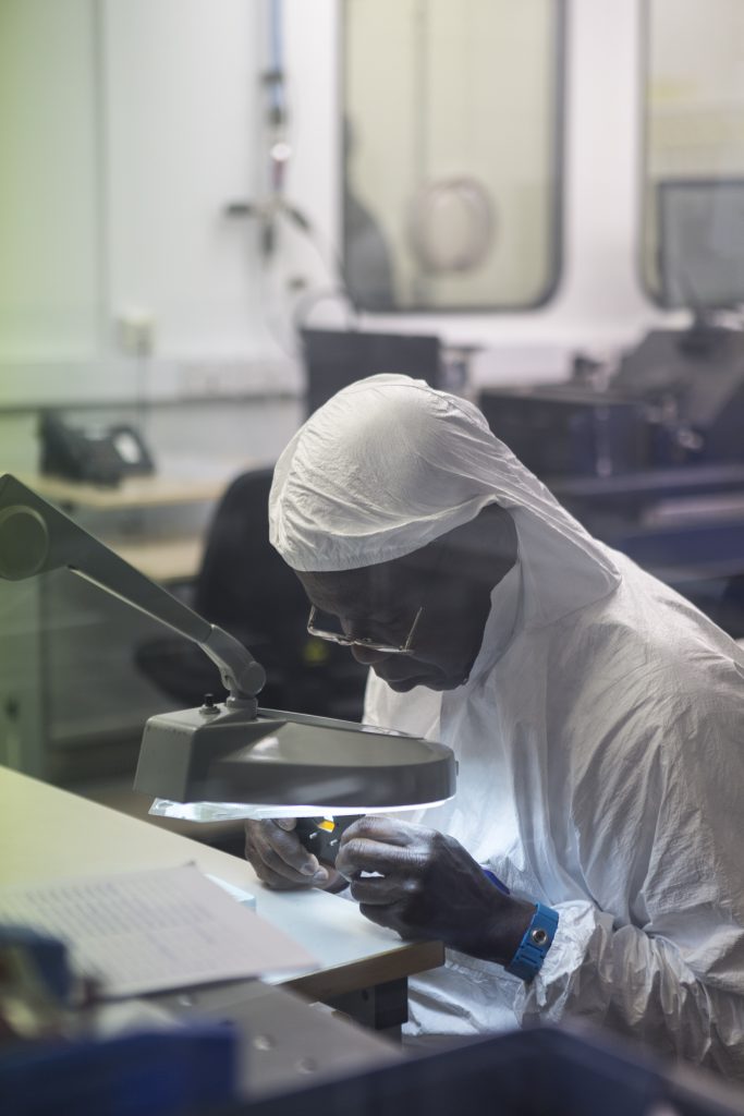 A man in a white protective suit uses a magnifying lens to help him assemble a camera.