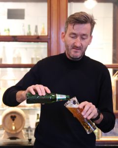 A man stands and pours a beer from a green bottle into a glass. He is blonde, with a beard, and is wearing a black turtleneck.