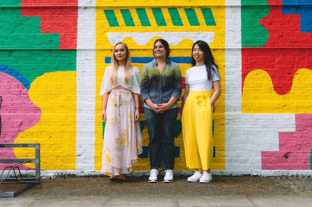 three women stand in front of colorful mural