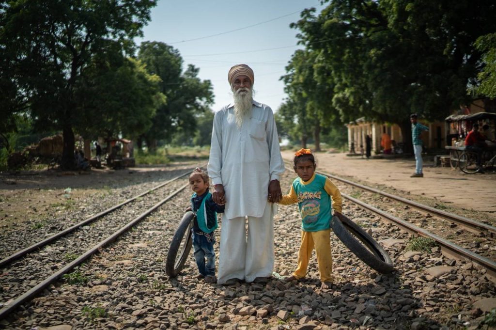 A grandfather stands near railroad tracks in rural India with two small children, each holding bicycle tires. 